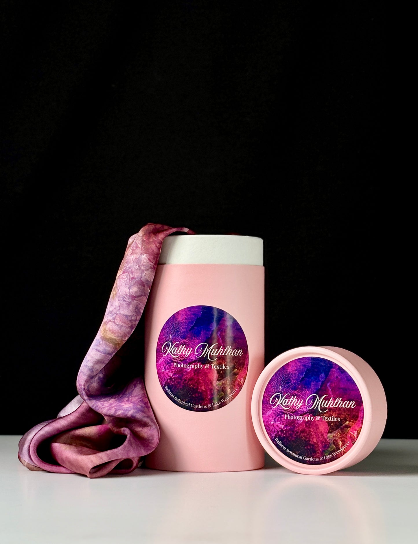 Pink cylindrical container with a purple and pink patterned label, accompanied by a matching pink lid. Coming out of the container is a Peony Satin Scarf cascading onto a white table, with a black background.