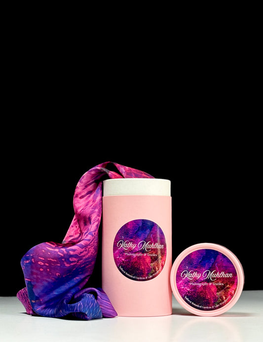 Pink cylindrical container with a purple and pink patterned label, accompanied by a matching pink lid. Coming out of the container is a Rhododendron Satin Scarf cascading onto a white table, with a black background.