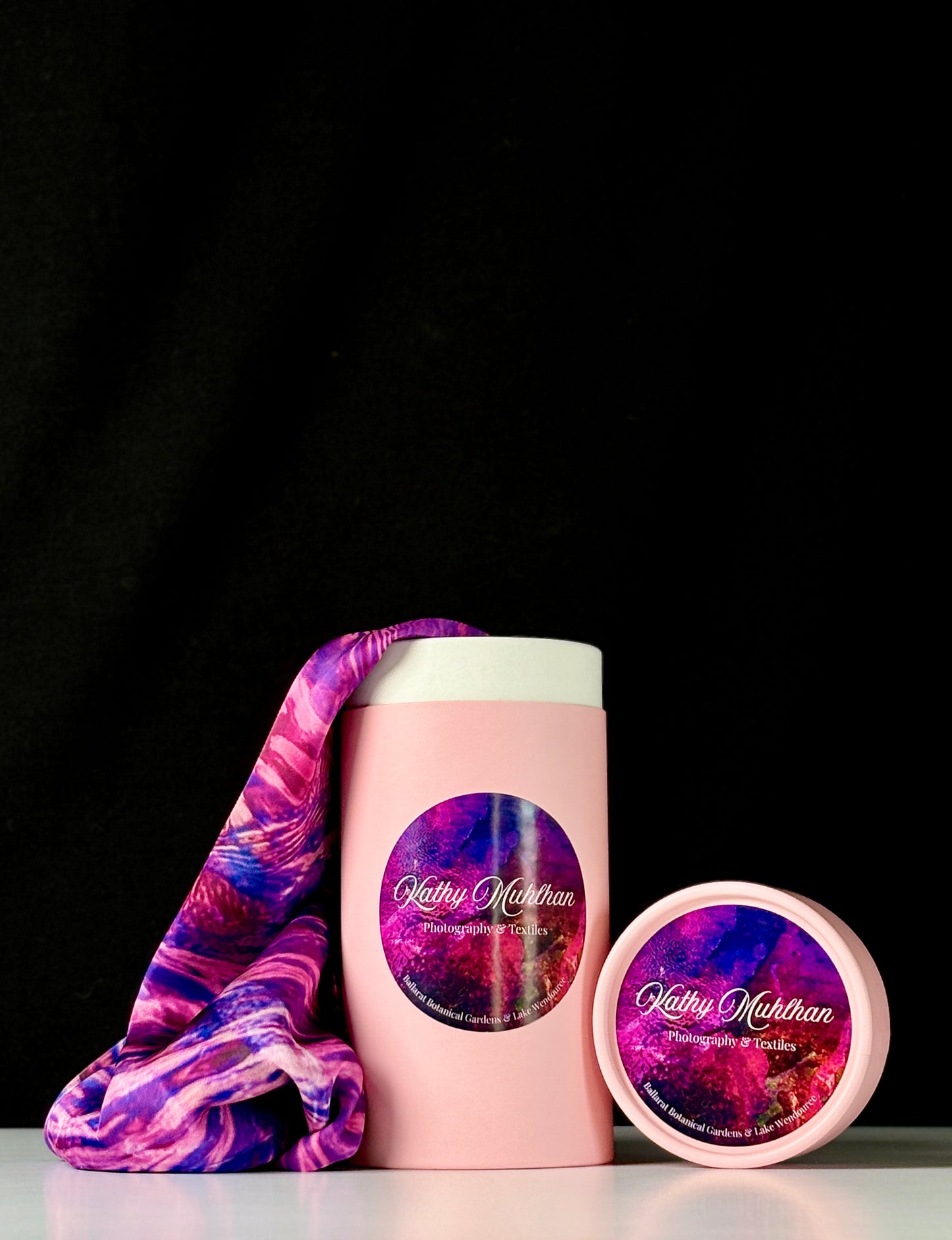 Pink cylindrical container with a purple and pink patterned label, accompanied by a matching pink lid. Coming out of the container is a Rosa Munstead Satin Scarf cascading onto a white table, with a black background.